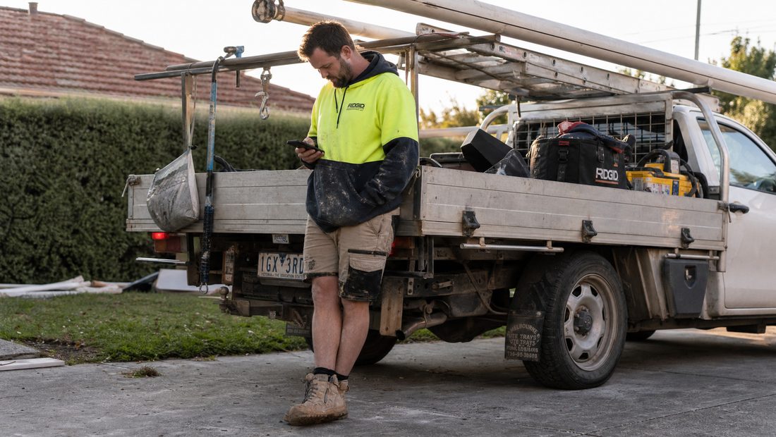 An Australian sole-trader tradie beside his ute comparing job-management apps on his phone
